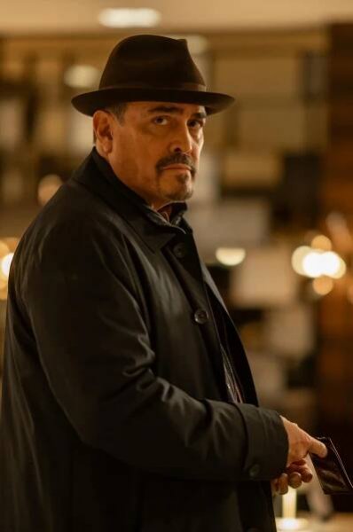 A man in a hat and dark coat stands in a lit room.