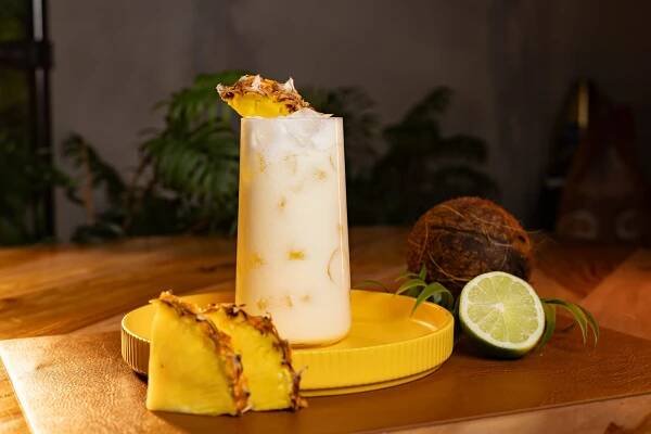 A side shot of a Coco Loco cocktail in a highball glass on a yellow tray placed on a wooden surface surrounded by a lime, a coconut and two pineapple slices