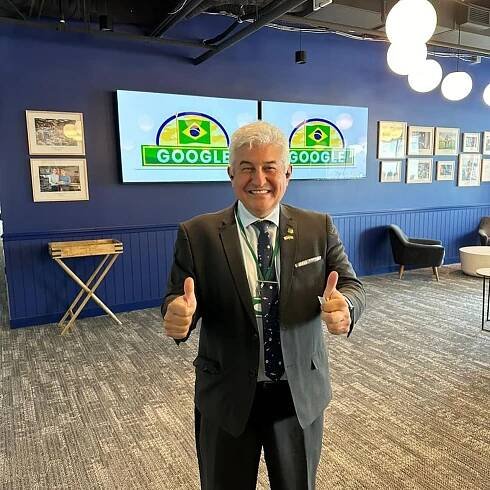 Senador Astronauta Marcos Pontes, que veste terno cinza escuro, camisa clara e gravata azul marinho com estampa espacial, sorri para a foto com os dois polegares levantados, fazendo joinha. Atrás, telas exibem logo do Google com referências à bandeira brasileira.