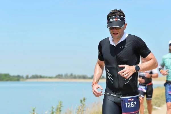 Guillermo Esteves running on a gravel trail, next to the Boulder Reservoir. He's wearing a black Roka trisuit, an Ironman visor, dark sunglasses, and a white ice rag around his neck. His race belt has a bib with the number 1267 on it.