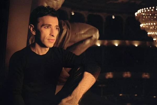 Portrait of a man in darkened theater. He's on the left of the frame with the sleeve of his black shirt rolled up. He gazes to the right; the lights in the theater glow but it's dim.