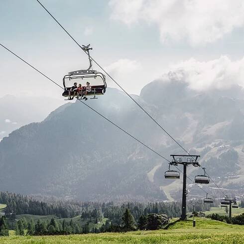 Drei Personen sitzen auf einer Seilbahnkabine über grüner Landschaft und bergiger Umgebung.