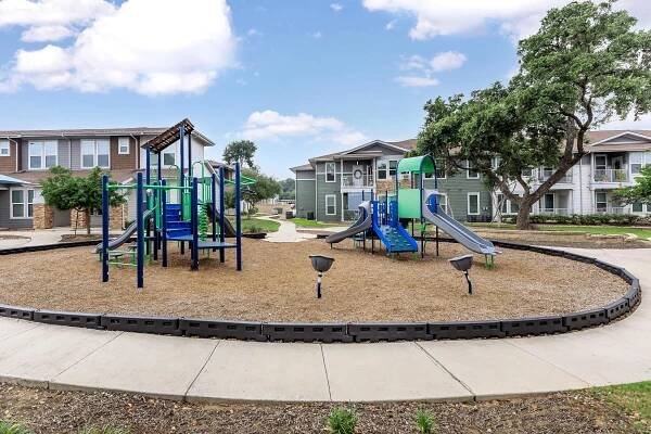 A playground area featuring two sets of play structures, including slides and climbing equipment, set on a bed of mulch. Surrounding the playground are pathways and residential buildings in the background, with greenery and blue skies contributing to a bright, inviting atmosphere.