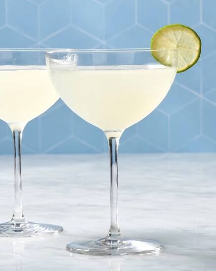 two classic daiquiri cocktails in coupe glasses, one with a lime garnish against a blue backdrop on a marble surface