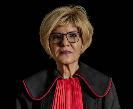 Das Porträtfoto einer blonden Frau mit schwarzer Brille, die in schwarz-roter Robe leicht herausfordernd in die Kamera blickt.