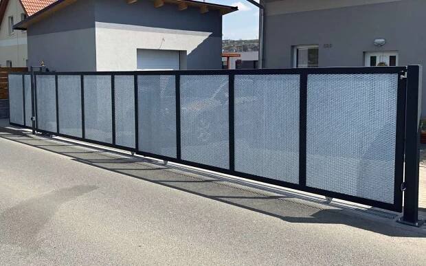Vjezdová posuvná brána na kolejnici teleskopická pro průjezd široký 6m s výplní tahokovu. Robustní rám šíře 70mm lakovaný v odstínu antracit, takohov žárově zinkovaný. Teleskopický mechanismus zkrátil potřebný prostor na zajetí bány na pouhých 3,6m. Realizace v Králově Dvoře u Berouna.