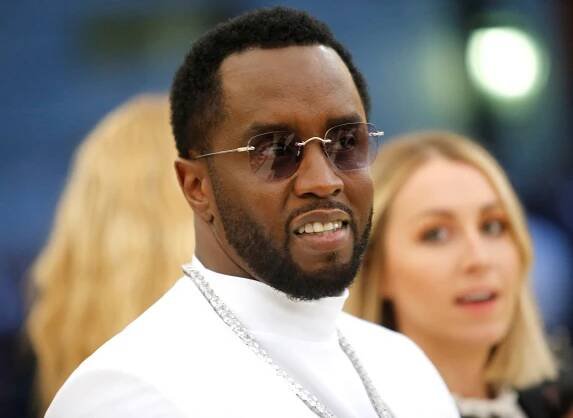 Sean Combs, dressed in white and in thin-frame sunglasses, slightly smiles.