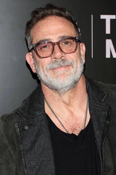 Jeffrey Dean Morgan with glasses and a beard smiles at an event, wearing a jacket over a shirt and layered necklaces