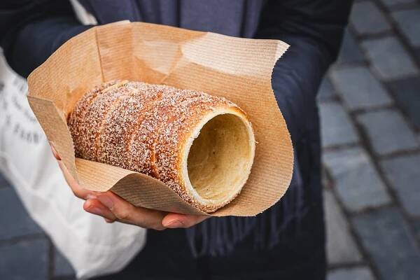Jin holds a Trdelník (chimney Cake) covered with sugar and cinnamon and wrapped in brown paper, which we bought at a Christmas Market in Prague