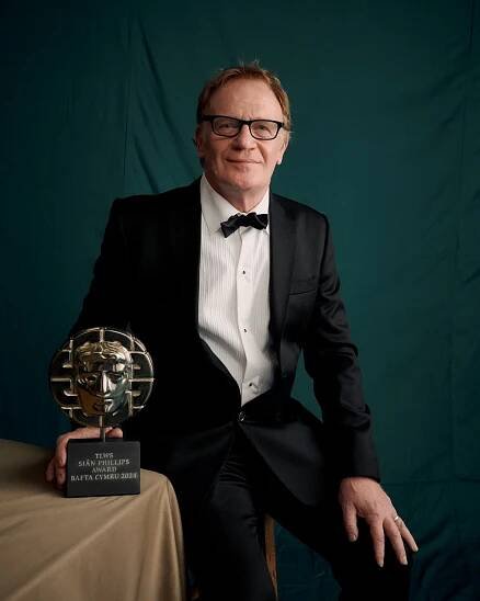 A white man in a suit with short grey hair and black-rimmed glasses. He is wearing a black tuxedo and bow tie and posing with his BAFTA.