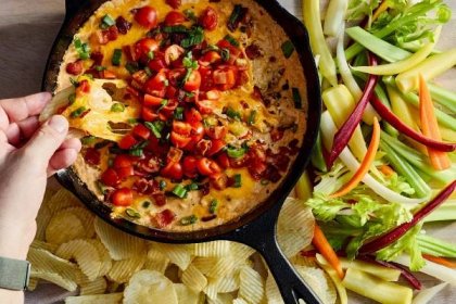 A hand dips a ruffled potato chip into a skillet full of bacon-Cheddar dip topped with tomatoes. An array of vegetables sits to the right of the skillet.