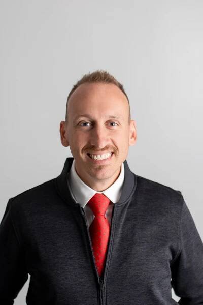 A professional headshot of Michael Cashman, a smiling man with short-cropped light brown hair and a mustache. He is wearing a white dress shirt, a red tie with small green accents, and a black zip-up jacket. The background is a plain light gray.