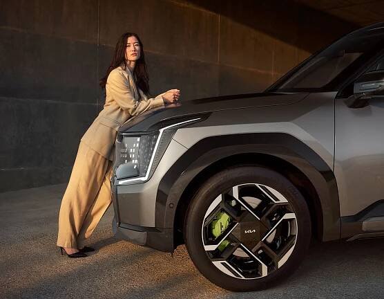 Kia EV9 GT, grau, Motorhaube Fahrerseite, Frau lehnt vorne am Fahrzeug