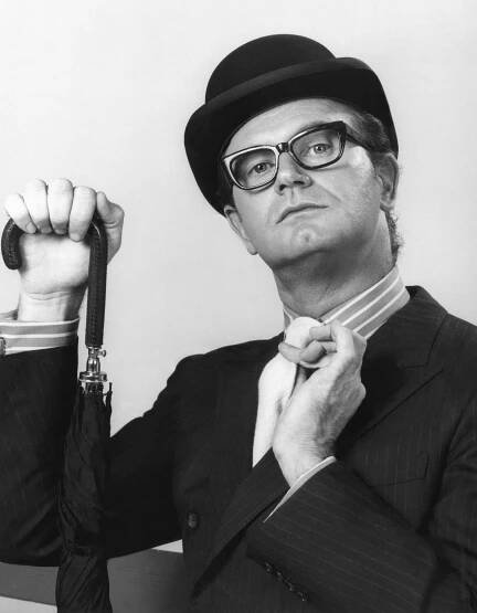 A man in a bowler hat and glasses holds an umbrella in one hand and puts the other hand up to his tightly knotted tie. 