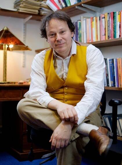 A color photograph of a seated man in a white shirt, khaki pants and a mustard-yellow vest. Behind the man we can see a desk and lamp and shelves with books. 