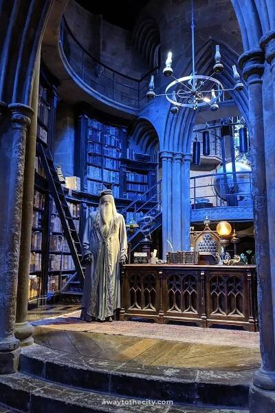 A detailed recreation of Albus Dumbledore's office at the Warner Bros. Studio Tour features towering bookshelves, a large ornate desk, and magical artifacts under a blue-toned light.