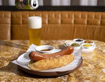 A hot dog placed next to a glass of beer on a wooden table at NYC restaurant Mischa.