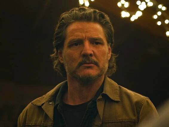A man with medium-length, swept-back gray and brown hair and a gray beard stands in an open space with fairy lights above him. He's wearing a brown jacket with dark button-up shirt and a t-shirt underneath.