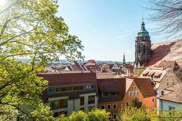 Eine malerische Aussicht auf eine europäische Stadt mit roten Ziegeldächern und einer markanten Kirche mit einem hohen Glockenturm. Grüne Bäume rahmen den Vordergrund und der Himmel ist klar und blau, was auf einen sonnigen Tag schließen lässt. Die Architektur ist traditionell und schafft eine malerische Kulisse.