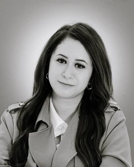 Professional black and white portrait photo of person with long dark hair wearing a beige trench coat and white blouse.
