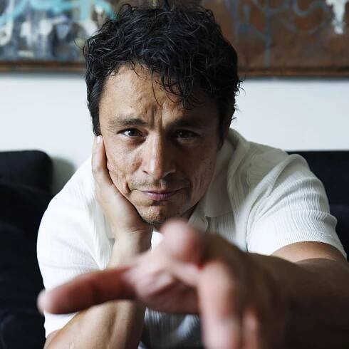 Jesse Garcia sits on a couch, reaching one hand out toward the camera