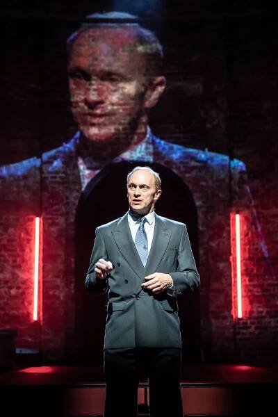 An actor in a gray double-breasted suit who looks a bit like President Vladimir Putin of Russia stands on a stage where his image is projected onto a brick wall behind him.