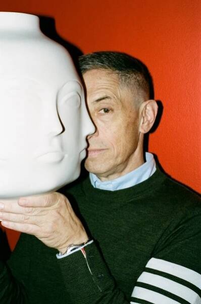 Against a red wall, a man in a dark sweater with a pale blue shirt collar peeking out, holds up a huge white ceramic head, with his own face pressed to one of the multiple faces of his creation.