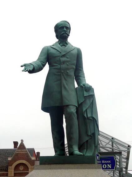 A statue depicting a mustachioed man holding an overcoat in his left hand with his right hand extended