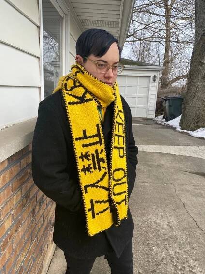 A whit man in all black winter wear wearing a bright yellow and black scarf.  The scarf has Japanese characters and english letters reading "KEEP OUT".  There is a brick halfwall and snow in the driveway behind him. 