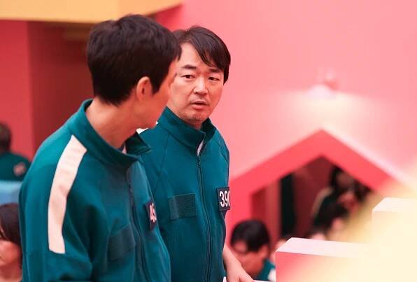 Two men in numbered teal tracksuits are talking, set in a colorful, geometric environment