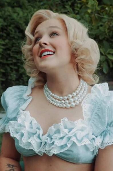 JoJo Siwa with a blonde, curly bob and wearing a ruffled crop top and pearl necklace, looks upward joyfully