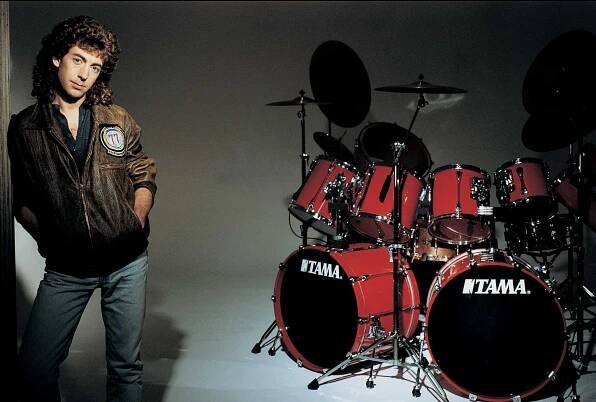 Drummer Simon Phillips leaning against a wall next to a red Tama drum kit