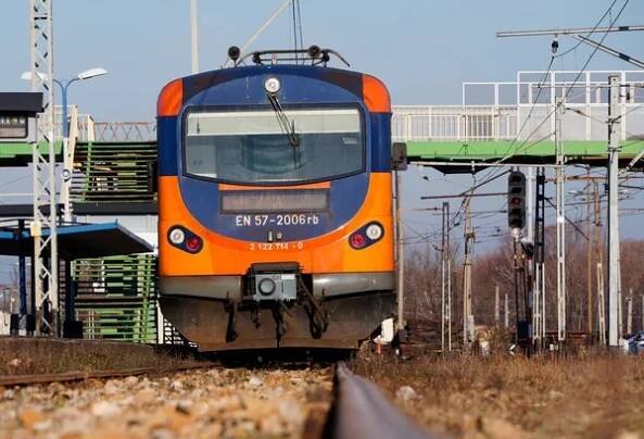Oranžový a modrý elektrický vlak na nádraží. Stopa, tyče a podpěry pro proudové tratě. — Stock obrázek