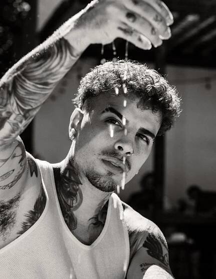 Rauw Alejandro, in a white tank top, drips water from the fingers of his tattooed right hand.