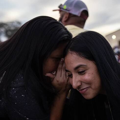 Gloria Cazares holds her hand up to her mouth and leans close to whisper into Kimberly Rubio's ear while outside. Rubio is smiling. Both have long dark hair and wear black T-shirts.