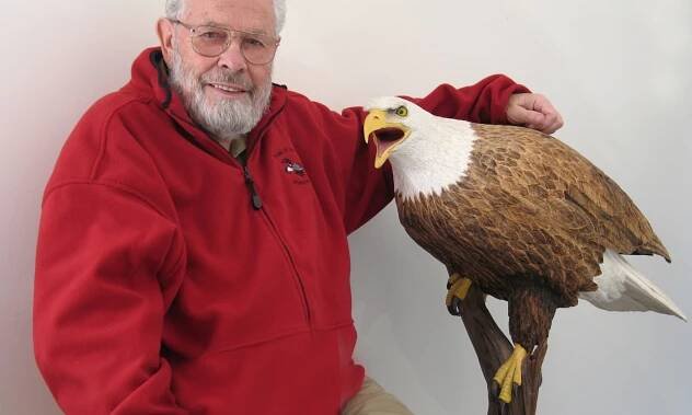 Bob Spear, Woodcarver, and his Bald Eagle carving: an elderly white mad with white hair and bear, wearing a red chamois shirt, faces the camera. His left arm rest on the back of a life-size bald eagle carved of wood.