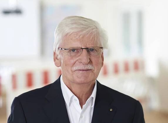 Älterer Mann mit weißem Haar, Brille und Anzug steht in einem modernen Büroraum