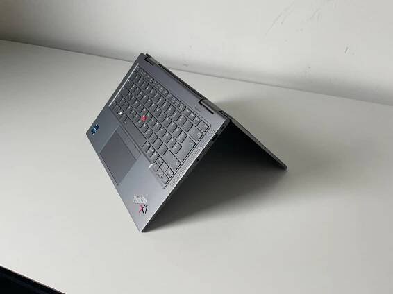 Ein Lenovo ThinkPad X1 Yoga Laptop in Zeltmodus auf einem weißen Tisch. Das Gerät hat ein graues Gehäuse, eine Tastatur mit rotem TrackPoint und das ThinkPad-X1-Logo auf der Handballenauflage. Der Bildschirm ist nach hinten geklappt, sodass das Gerät wie ein umgedrehtes 