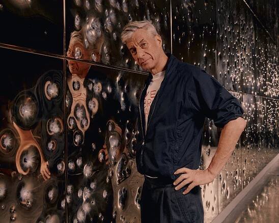 Maurizio Cattelan, with silver hair, in black pants and a black shirt, posing for a portrait at Gagosian with part of his show titled “Sunday.” It consists of steel panels plated with gold and riddled with bullets from various weapons.