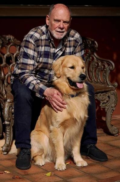Gordon Eubanks Jr. in a plaid shirt sits on a bench, smiling with his arm around a golden retriever.