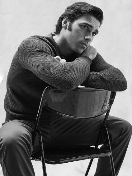 Giuseppe Maggio - A man sits on a backward-facing chair, resting his arms and chin on the chair’s back. He appears thoughtful, gazing slightly upward. The image is in black and white.