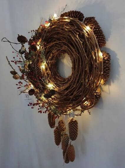 a wreath with pine cones and lights hanging from the side on a white wall behind it