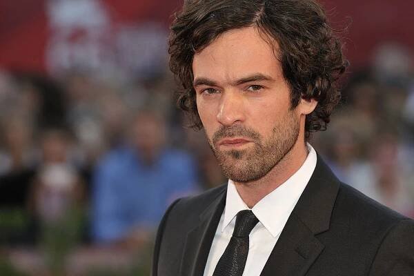 Young bearded actor wearing a white shirt and a black suit.