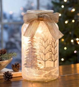 a glass jar filled with snow and pine trees on top of a wooden table next to a christmas tree