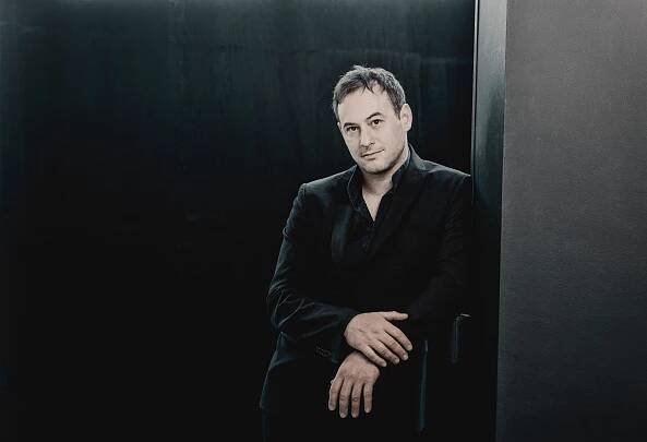 A white man wearing a black suit leaning against a grey wall