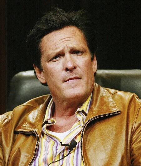 A close-up photo of him in a brown leather jacket sitting in a chair while looking at the camera with knitted brow and a serious expression. 