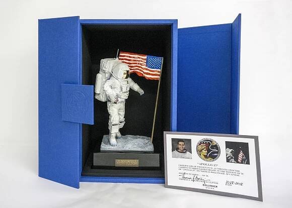 Image of the final sculpture maquette of Harrison Schmitt in space suit next to an American Flag. The maquette is placed within a bright blue box, alongside a certificate of  authenticity. 