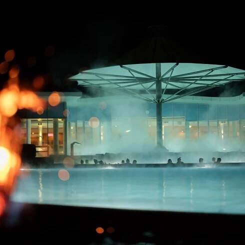 Nachtaufnahme vom Außenbereich in die beleuchtete Therme, mit Fackellicht im Vordergrund