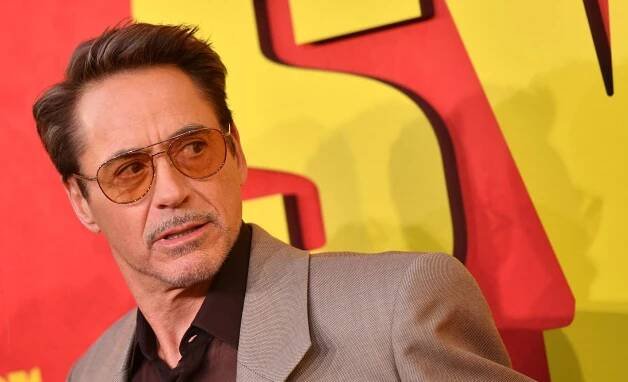 Robert Downey Jr., with a goatee and wearing tinted, aviator glasses, stands in front of a red and yellow backdrop.