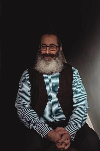 Seth Barrett Tillman posing for a portrait while seated. He has a long gray beard and wears a black vest over a striped shirt.